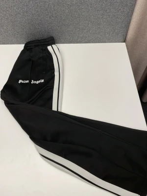 Palm Angels svarta trackpants S - Svarta trackpants från Palm Angels med vita sidoränder och logga på vänster ben. Byxorna har resår i midjan, dragkedja vid benslut och är gjorda i 100% polyester för en sportig vibe. Perfekta för en streetwear-look. Skriv för frågor!