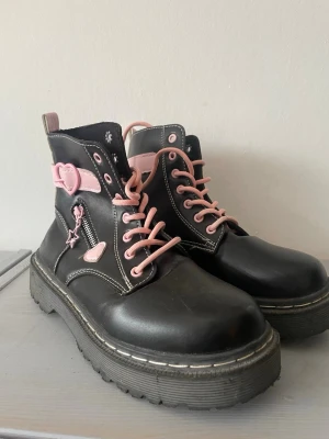 Svarta boots med rosa detaljer - Säljer ett par chunky svarta boots med rosa snörning och stjärnberlock vid dragkedjan. Skorna har grov sula, vita sömmar och rund tå. Perfekta för dig som vill sticka ut med söta detaljer och edgy vibe.
