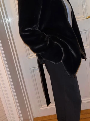 Daisy Grace – svart fuskpälsjacka med skärp, XS, ny med prislapp - Elegant svart fuskpälsjacka från Daisy Grace. Mjuk och följsam kvalitet med stilrena, blanka detaljer längs framkanten samt matchande knytskärp som markerar midjan. Rak modell som kan bäras både öppen och knuten.  Jackan passar lika bra till vardag som till festligare tillfällen. ✔ Nypris: 1 400 kr ✔ Oanvänd ✔ Prislapp kvar