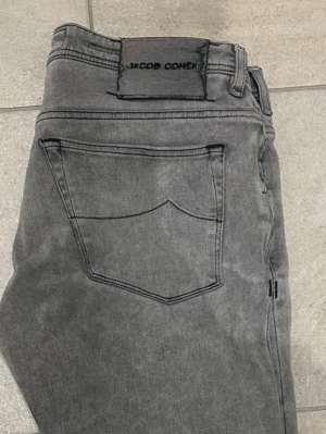 Grå jeans från Jacob Cohën - Snygga grå jeans från Jacob Cohën med klassisk femficksdesign och raka ben. Jeansen har en diskret tvättad look och svart patch med broderad logga bak i midjan. Tillverkade i mjukt bomullsmaterial för skön passform.