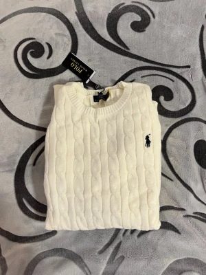 Vit kabelstickad tröja Polo Ralph Lauren - Säljer en vit kabelstickad tröja från Polo Ralph Lauren i 100% bomull. Klassisk rund halsringning, ribbade muddar och ikoniska loggan broderad på bröstet. Perfekt till lager på lager och riktigt snygg till jeans eller kjol.