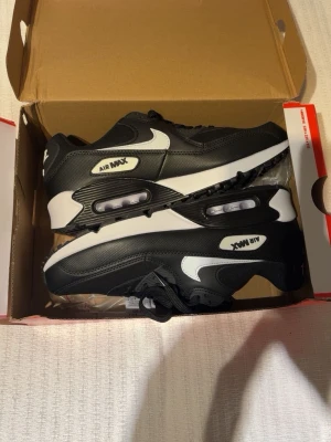 Nike Air Max 90 svart/vit sneakers - Nike Air Max 90 sneakers i svart och vitt med klassisk synlig Air-enhet i sulan. Skorna har en vit swoosh på sidan, svarta snören och detaljer i syntetmaterial och skinn. Perfekt för dig som gillar stilrena och sportiga sneakers. Helt nya, box ingår!!