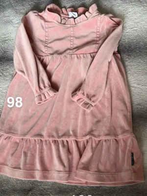 Rosa volangklänning i velour - Supersöt rosa klänning från Polarn o pyret i mjuk velour. Klänningen har långa ärmar, volangdetaljer vid ärmslut och nederkant samt en liten volangkrage. Perfekt för dig som gillar gulliga och bekväma plagg!🌸 Fint använt skick. Storlek 98. 