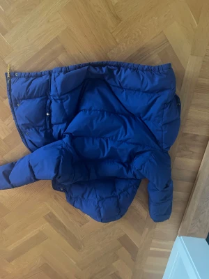 Blå dunjacka från Polo Ralph Lauren - Säljer en blå dunjacka från Polo Ralph Lauren med röd insida och huva. Jackan har silverfärgade knappar, två fickor med knappstängning och elastisk nederkant. Perfekt för kalla dagar och har en klassisk streetstil med snygg färgkombination.