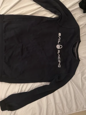 Svart Sail Racing sweatshirt - Säljer en svart sweatshirt från Sail Racing med vit logga tryckt på bröstet och en mindre logga i nacken. Tröjan har rund halsringning och långa ärmar. Perfekt för dig som gillar stilren och sportig design. Knappt använd