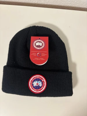 Svart ribbstickad mössa Canada Goose - Säljer en svart ribbstickad mössa från Canada Goose med klassisk Arctic Program-patch framtill. Mössan är i mjukt material och har vikt kant för extra stilpoäng. Perfekt för kalla dagar och snygg till alla vinteroutfits.