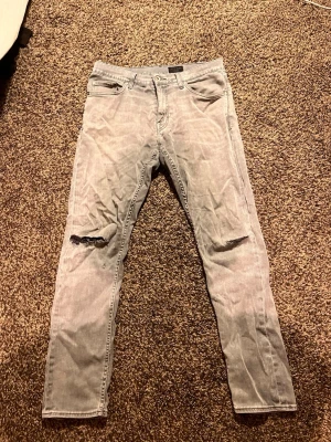 Grå jeans Tiger of Sweden Pistolero 31/32 - Säljer ett par grå jeans från Tiger of Sweden i modellen Pistolero. Jeansen har skinny passform, slitningar vid knäna och klassisk femficksdesign. Materialet är jeans och färgen är ljusgrå, perfekt för dig som gillar en clean men edgy stil.