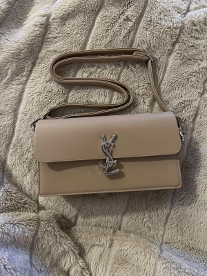 Beige axelväska från YSL - Snygg beige axelväska från YSL med klassisk silverfärgad logga framtill. Väskan har en stilren, rektangulär form och justerbar axelrem. Perfekt för dig som gillar en clean och lyxig look. Tillverkad i slätt skinn som känns exklusivt och tidlöst.