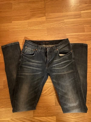 Mörkblå jeans från Nudie Jeans - Säljer ett par mörkblå jeans från Nudie Jeans med snygg slitning och klassisk femficksmodell. Jeansen har raka ben och normal passform, tillverkade i slitstarkt denimtyg av bomull. Märkespatch bak i midjan och diskret broderad detalj på bakfickorna.