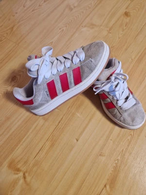 Adidas Campus sneakers beige och röd - Säljer ett par Adidas Campus sneakers i beige mocka med röda detaljer och snörning. Skorna har de klassiska tre ränderna i rött på sidorna och en vit sula med gummisula undertill. Perfekt för dig som gillar retrostil och vill sticka ut med färg.