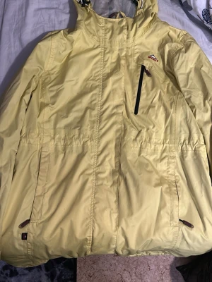 Gul tun jacka från McKinley - Ljusgul vindjacka från McKinley med huva och flera fickor med dragkedja. Jackan har en sportig look och är tillverkad i lätt syntetmaterial. Märkt med 'exodus 5000' på ärmen och har justerbara detaljer vid ärmslut och midja.