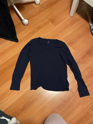 Mörkblå långärmad topp med rosett - En enkel och stilren mörkblå långärmad topp från L.O.G.G. i mjuk bomull. Toppen har en liten söt rosett framtill vid halsringningen och en klassisk, rak passform. Perfekt att matcha med jeans eller kjol för en clean look.