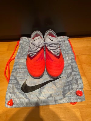 Nike fotbollsskor röd/vit med snörning - Säljer ett par snygga Nike fotbollsskor i rött och vitt med svarta detaljer. Skorna har snörning, rund tå och platt sula med fasta dobbar för gräsplan. Ovandelen är i syntetmaterial och designen är riktigt sportig. Perfekt för dig som vill sticka ut på planen!