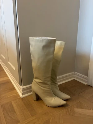 Beiga höga boots med klack - Säljer ett par höga boots i beige skinnimitation med spetsig tå och bred klack. Skorna har dragkedja på insidan för enkel påtagning och en stilren, minimalistisk look. Perfekta för dig som vill sticka ut med ett par statement boots.