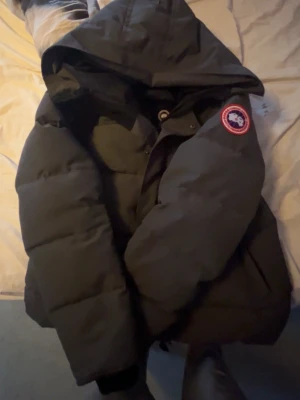 Svart pufferjacka från Canada Goose M - Säljer en svart pufferjacka från Canada Goose i storlek M. Jackan har en stor huva, klassisk Canada Goose-logga på ärmen och stängs med både dragkedja och knappar. Perfekt för kalla dagar och riktigt snygg till streetwear.