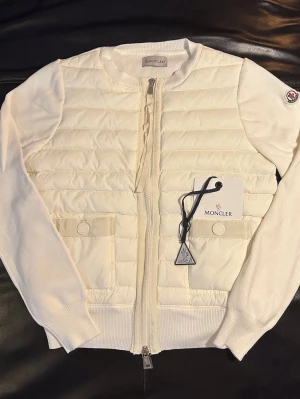 Moncler Cardigan  - Säljer en snygg vit quiltad jacka från Moncler med ribbade stickade ärmar och två fickor framtill. Jackan har dragkedja hela vägen och Moncler-logga på ärmen. Perfekt för dig som vill ha en stilren och exklusiv look.