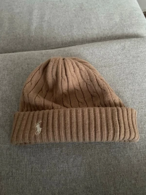 Ralph Lauren mössa - Säljer en beige stickad mössa från Polo Ralph Lauren med klassisk kabelstickning och vikt kant. Mössan har den ikoniska broderade loggan framtill och är tillverkad i 80% ull och 20% nylon. Perfekt accessoar för kyliga dagar.