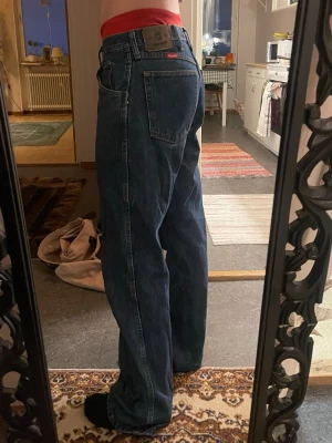 Wrangler blå raka jeans - Säljer ett par klassiska blå jeans från Wrangler med rak passform och normal midja. Storlek 34 x 32. Jeansen har fem fickor, tydliga sömmar och Wrangler-lapp bak i midjan. Materialet är robust denim i bomull och färgen är mörkblå. Perfekta för en avslappnad och tidlös look.