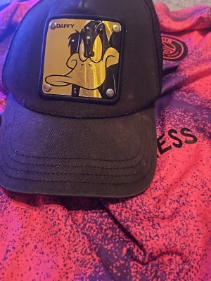 Svart Daffy Duck keps från Capslab - Säljer en svart keps från Capslab med Daffy Duck-platta i guld framtill. Kepsen har mesh på sidorna och bak, samt broderad Daffy Duck-text. Officiell Looney Tunes-produkt, perfekt för dig som gillar tecknade klassiker och streetstyle.