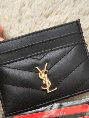 Svart quiltad plånbok i skinn - Säljer en stilren svart plånbok med quiltad design och guldfärgad logga framtill. Perfekt för kort och småsaker, tillverkad i mjukt skinn och har flera fack för enkel organisering.