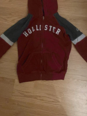 HOLLISTER  KOFTA -  Hoodien har en djup röd färg på kroppen och luvan, med grå ärmar. Den har en dragkedja fram och en känguruficka. På bröstet står det "HOLLISTER" i vita bokstäver. Ärmarna har en grå färg, med en ljusare grå och vit rand nära handleden. 