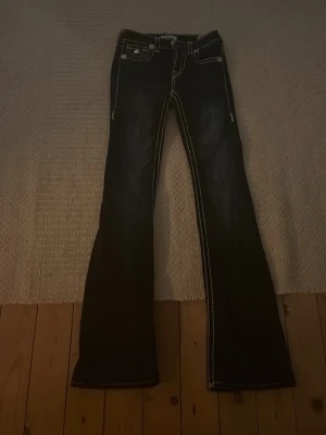 Blåa bootcut jeans med vita sömmar - Säljer ett par svarta jeans från True Religion med tydliga vita kontrastsömmar och bootcut-modell. Jeansen har klassiska fickor fram och bak, samt dekorativa detaljer på bakfickorna. Materialet är jeans och passformen är normal. Kostar 2400 i orginalpris 