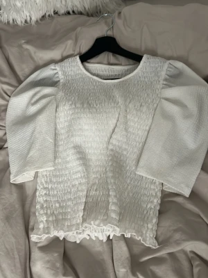 Vit blus med puffärm och smock - Supersöt vit blus med puffiga trekvartsärmar och smockad kropp. Blusen har rund halsringning och volangkant nertill som ger en romantisk vibe. Perfekt att styla med jeans eller kjol för en trendig look.