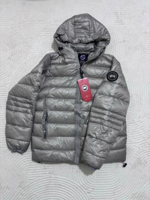 Grå dunjacka från Canada Goose - Säljer en grå dunjacka från Canada Goose med huva och dragkedja framtill. Jackan har quiltad design, två sidofickor med dragkedja och en snygg patch på ärmen. 