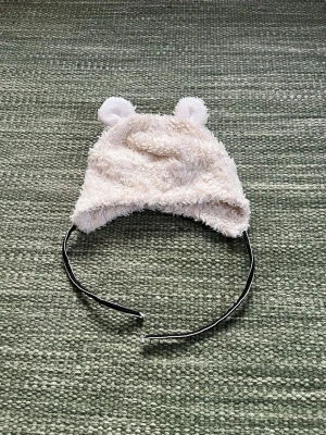 Vit fluffig barnmössa med öron - Supersöt vit fluffig mössa från Åhléns med små öron på toppen. Mössan är gjord i mjukt syntetmaterial och har ett gulligt nallebjörnsutseende. Perfekt för att hålla värmen och addera lite lekfull stil till din outfit.