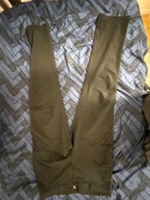 Svarta täckbyxor från Trekmates, stl S - Säljer ett par svarta täckbyxor från Trekmates i storlek S. Byxorna har raka ben, dragkedjor vid fickorna och är gjorda i ett slitstarkt syntetmaterial. Perfekta för utomhusaktiviteter och äventyr. Snygg och funktionell design med diskret logga på benet.