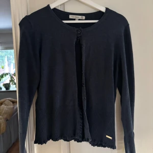 Mörkblå kofta från Flash Woman - Mörkblå kofta från Flash Woman med rund halsringning och knappar framtill. Koftan har långa ärmar, ribbade muddar och en liten volangkant nedtill. Perfekt för lager-på-lager och enkel att matcha med olika outfits.