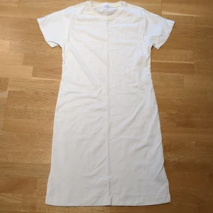 Vit t-shirtklänning från Filippa K - Vit t-shirtklänning från Filippa K i ekologisk bomull. Enkel och stilren design med rund halsringning och korta ärmar. Rak passform och knälång modell, perfekt för en clean och avslappnad look.