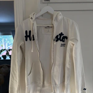 Vit hoodie från Hollister med dragkedja - Vit hoodie från Hollister med broderad mörkblå logga och text på bröstet. Tröjan har huva med snörning, två fickor fram och dragkedja hela vägen. Dragkedjan funkar ibland så om man kan fixa de så funkar den. Perfekt för chill dagar.