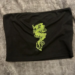 Svart tubtopp med draktryck från SHEIN - Cool svart tubtopp från SHEIN i storlek XS med ett gult drakmotiv framtill. Toppen är axelbandslös och har en enkel, tight passform som sitter snyggt. Perfekt för dig som vill sticka ut med en edgy vibe.