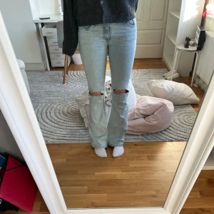 Ljusblå bootcut jeans med hål på knäna - Säljer ett par ljusblå jeans med bootcut-modell och slitna hål på båda knäna. Jeansen har klassisk femficksdesign och en snygg broderad detalj på bakfickorna. Perfekta för dig som gillar en avslappnad och trendig stil.