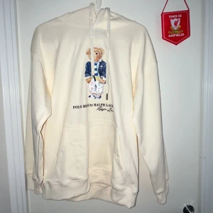 Polo Bear hoodie från Ralph Lauren - Krämvit hoodie från Ralph Lauren med Polo Bear-tryck framtill. Tröjan har huva med snörning, känguruficka och långa ärmar. Materialet känns mjukt och skönt, perfekt för chill dagar. Klassisk och stilren design med ikonisk björn.