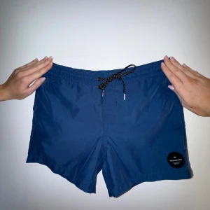 Blå Quiksilver badshorts med snörning - Snygga marinblå badshorts från Quiksilver med elastisk midja och svartvit snörning framtill. Shortsen har en rund svart patch med logga på benet och en bakficka med Quiksilver-märke. Perfekta för strand eller poolhäng!