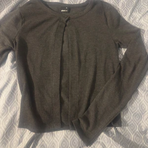 Mörkgrå kofta från Gina Tricot - En stilren mörkgrå kofta från Gina Tricot med rund halsringning och långa ärmar. Koftan har en enkel design utan synliga knappar och är tillverkad i ett mjukt, stickat material som passar perfekt till lager-på-lager-stil.