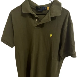 Olivgrön pikétröja från Polo Ralph Lauren - Snygg olivgrön pikétröja från Polo Ralph Lauren i custom slim fit. Klassisk krage, korta ärmar och gul broderad logga på bröstet. Tillverkad i bomull, perfekt för en clean och stilren look.