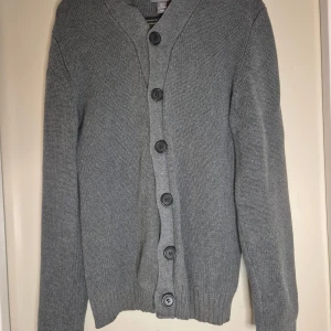 Grå stickad kofta från H&M - Mysig grå stickad kofta från H&M med stora svarta knappar framtill. Koftan har långärmad passform, ribbade muddar och en klassisk v-ringning. Perfekt för lager-på-lager och chill dagar.