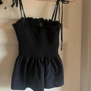 Svart smockad linnetopp från Loness - Supersöt svart linnetopp från Loness med smockad överdel och volangkant upptill. Toppen har tunna axelband som knyts och en lätt utställd nederdel. Perfekt för en trendig och avslappnad look.