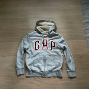 Grå fur GAP hoodie - Mysig grå hoodie från GAP med röd-vit logga framtill. Tröjan har dragkedja, huva och är fodrad med fluffig beige fleece på insidan. Perfekt för kyliga dagar och har två fickor framtill. Klassisk och enkel stil med långa ärmar.