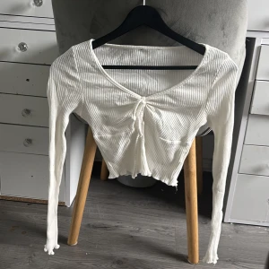 Vit ribbad croppad topp med snörning - Säljer en vit ribbad långärmad topp med croppad passform och snörning framtill. Toppen har v-ringning och lätt vågig kant nertill för en trendig look. Perfekt för dig som gillar stilrena och enkla plagg med en twist.