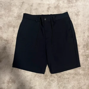 Svarta shorts med snörning - Snygga svarta shorts med elastisk midja och snörning framtill. Klassisk rak passform med sidofickor och bakfickor med knapp. Perfekta för sommardagar och enkel att matcha med allt. Stilrena och bekväma för en avslappnad look.