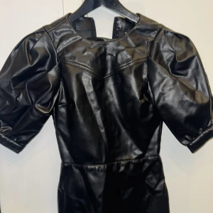 Svart klänning i skinnimitation - Cool svart klänning i skinnimitation med puffiga korta ärmar och rund halsringning. Snygg sömdetalj över bröstet och markerad midja. Perfekt för dig som vill sticka ut med en edgy look.