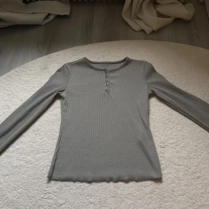 Ribbad grå långärmad topp med knappar - Snygg ribbad grå långärmad topp med rund halsringning och sex knappar framtill. Figurnära passform och mjukt material som känns skönt mot huden. Perfekt att styla med jeans eller kjol för en clean look. Helt ny!! 