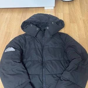Svart pufferjacka från Superdry - Svart pufferjacka från Superdry med huva och dragkedja framtill. Jackan har broderad logga på ärmen och ryggen samt quiltad design. Perfekt för kalla dagar och har en clean, jacka är S men passar även M, sportig look.perfekt nu till kommande kalla vädret:)