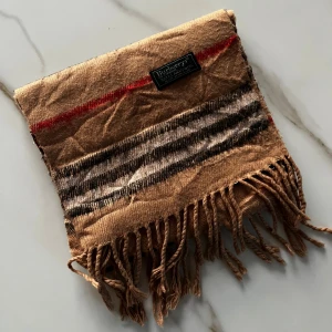 Burberry halsduk beige - Liten beige halsduk från Burberry med svarta, vita och röda ränder. Gjord i 100% lammull och har snygga fransar i ändarna. Perfekt accessoar för att lyfta din outfit med en tidlös brittisk touch. Säljs billigt eftersom att det är en mindre och annan modellen en den vanliga!