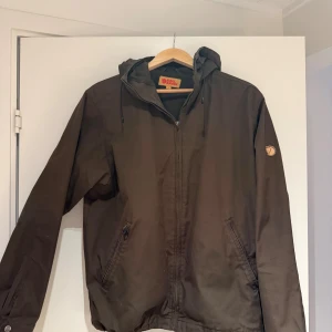 Brun vindjacka från Fjällräven M - Säljer en brun vindjacka från Fjällräven i G-1000-material. Jackan har huva, dragkedja framtill, två fickor med dragkedja och klassisk logga på ärmen. Perfekt för dig som gillar stilren och funktionell design.