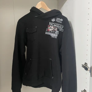 Svart hoodie Geographical Norway - Svart hoodie från Geographical Norway med tryck och broderade detaljer fram, bak och på ärmen. Stor norsk flagga på ryggen, bröstficka med knapp och känguruficka. Mjuk insida och huva med grått foder.